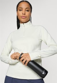 Woman with braided hair wearing a white long-sleeve athletic top opening a black Nike water bottle against a plain background.