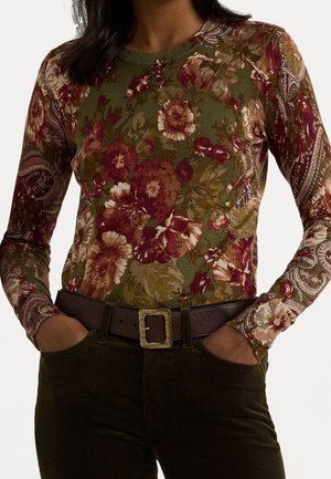 Osoba ubrana w długi rękaw z kwiatowym wzorem w odcieniach zieleni, czerwieni i beżu, z rękami w kieszeniach ciemnozielonych spodni sztruksowych.