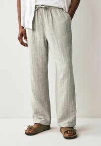 Pantaloni din in gri deschis cu o croială lejeră, având un brâu elastic și o textură subtilă, asortați cu sandale maronii.