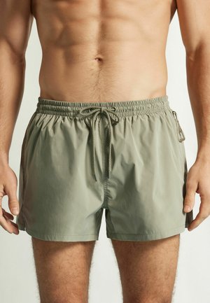 Mann trägt hellgrüne Badehose mit elastischem Bund, vorderer Kordel und seitlicher Reißverschlusstasche, steht vor einem einfarbigen Hintergrund.