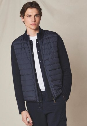 Junger Mann trägt eine marineblaue gesteppte Weste über einem marineblauen Cardigan und einem weißen T-Shirt, steht vor einem schlichten beigen Hintergrund.