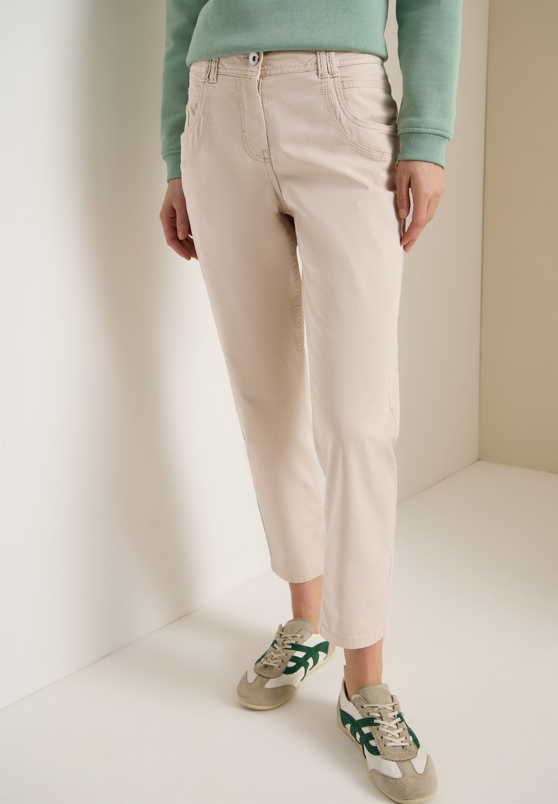 Personne portant un pantalon beige court, des baskets vertes et blanches, et un haut à manches longues vert, se tenant contre un mur bicolore.