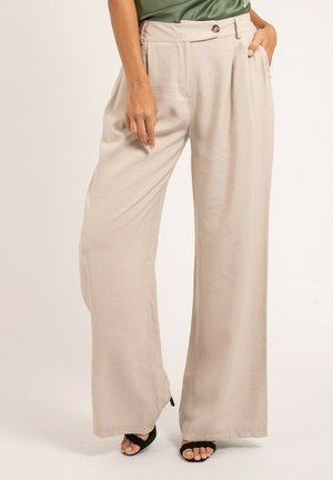 Femme portant un pantalon large beige à taille boutonnée et des talons ouverts noirs, debout avec une main dans la poche devant un fond uni.