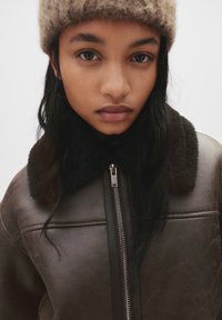 Jonge vrouw met lang donker haar, die een beige gebreide muts en een bruin leren jack met een shearling kraag en ritssluiting aan de voorkant draagt.