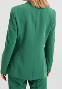 Zielona, dopasowana marynarka wykonana z gładkiej tkaniny. Charakteryzuje się strukturalnym krojem, klapą z wycięciem oraz pojedynczym rozcięciem z tyłu, z czystymi liniami.