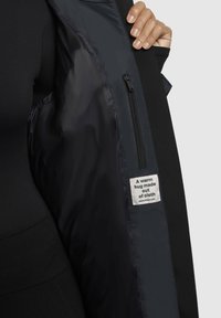 Chaqueta negra con un forro interior suave y brillante. Presenta un bolsillo con cremallera y una etiqueta que dice "Un abrazo cálido hecho de tela."