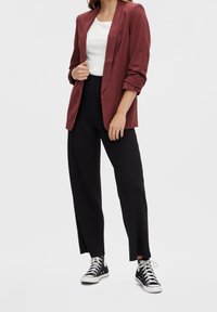 Femme portant un blazer bordeaux par-dessus un haut blanc, un pantalon large noir et des baskets montantes noires, debout devant un fond uni.