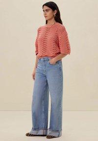 Koraal kleurige gebreide top met een losse, luchtige uitstraling, gecombineerd met wijde lichtblauwe jeans en zwart-bruine luipaardprint schoenen.