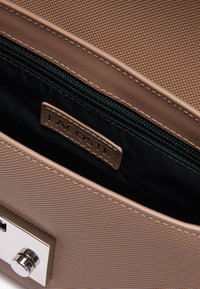 Borsa beige con esterno testurizzato, hardware in argento e interno nero con zip. Presenta un'etichetta con il logo Lacoste all'interno.