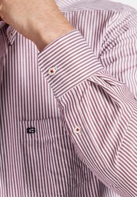 Chemise rayée rouge et blanche avec un col boutonné, des boutons blancs avec des accents rouges, et une poche poitrine gauche avec un logo brodé.