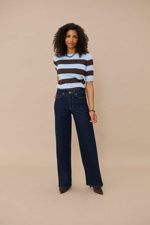 Femme aux cheveux bouclés porte un t-shirt à manches courtes rayé bleu et marron, un jean large foncé, des chaussures marron ainsi qu'un collier et un bracelet de perles.