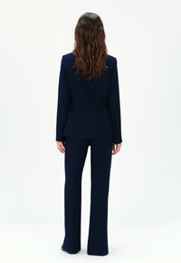Abito blu navy su misura con giacca aderente e pantaloni a gamba larga, caratterizzato da una texture liscia e dettagli minimi, visto da dietro.
