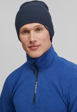 Navy knit beanie with ribbed texture, worn by a person in a bright blue fleece jacket with half-zip, featuring a logo on the collar.