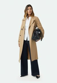 Beige Trenchcoat mit großen Knöpfen, breiten Revers und lockerer Passform, kombiniert mit einem weißen Hemd mit Kragen, dunklen weiten Hosen und schwarzen High Heels.