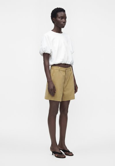 Γυναίκα που στέκεται, φοράει λευκό cropped top με φουσκωτά μανίκια, μπεζ σορτς με ψηλή μέση και μαύρα σανδάλια με τακούνι σε απλό λευκό φόντο.