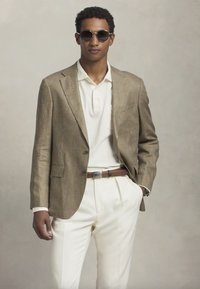 Sacou căptușit maro, cu carouri, purtat peste un tricou polo alb, asortat cu pantaloni de culoare crem și o curea maro. Modelul poartă ochelari de soare negri.