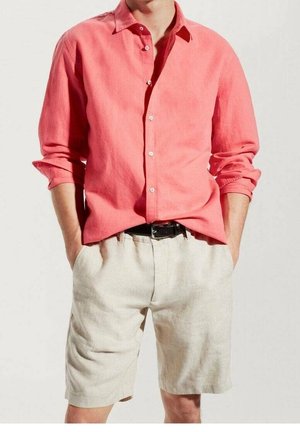 Uomo che indossa una camicia a maniche lunghe color corallo con bottoni e maniche rimboccate e pantaloncini beige con cintura nera, con le mani in tasca, in piedi.