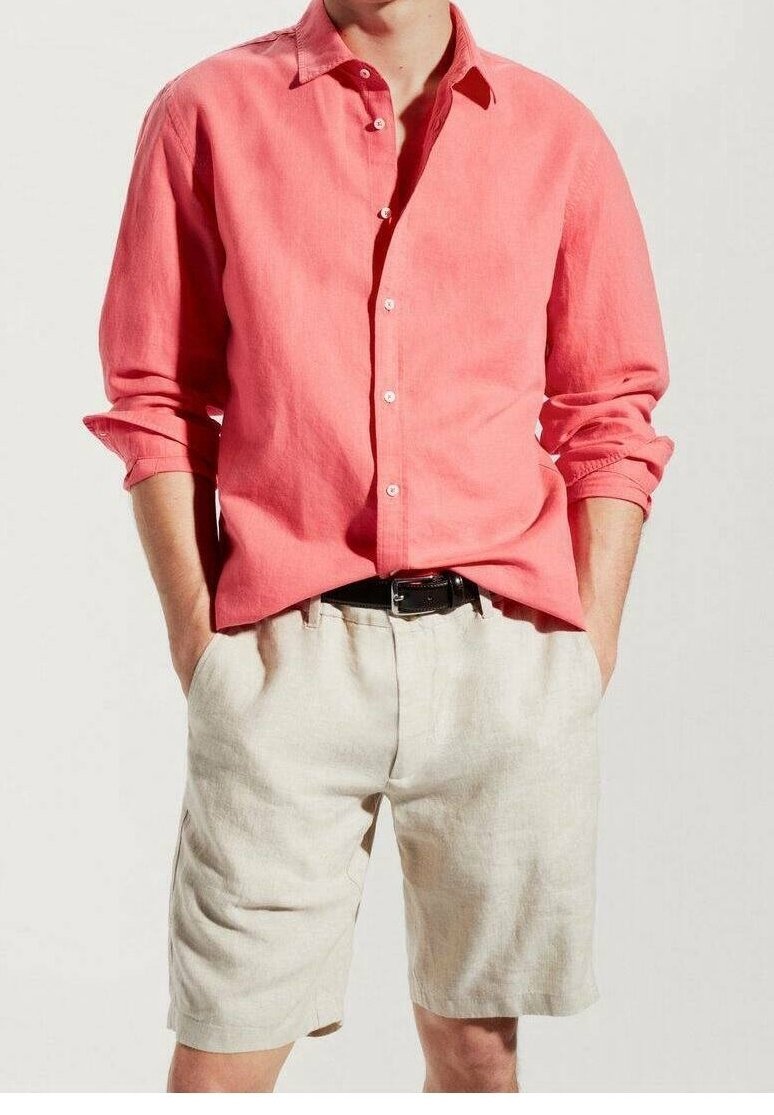 Homme portant une chemise corail à manches longues avec les manches retroussées et un short beige avec une ceinture noire, les mains dans les poches, debout.