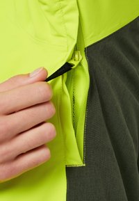 Main tenant un curseur de fermeture éclair noir dézippant partiellement une veste en tissu jaune vif et gris foncé.