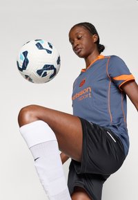 Grå sporttröja med orange detaljer, svarta shorts och vita knähöga strumpor. En mönstrad fotboll balanseras på ett uppdraget knä.