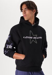 Sort hættetrøje med frontlogoet "LOVE SCARS" i rhinsten, stjernemotiver i lilla og hvid på ærmerne, kengurulomme, afslappet pasform.