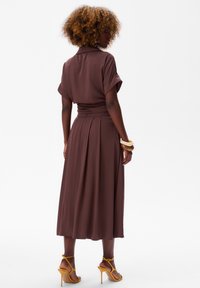 Bruine midi-jurk met korte mouwen, een ceintuurlijke taille en een vloeiende rok. Glad materiaal, plooien aan de achterkant, en gecombineerd met gele haksandalen.