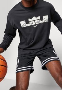 Maglietta nera a maniche lunghe con logo bianco geometrico e branding Nike, abbinata a pantaloni corti neri perforati con strisce bianche sui lati.
