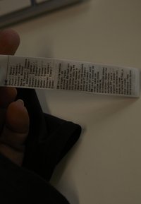 Hånd holder et tøjmærke, der viser fiberindhold og vaskeanvisninger på flere sprog på et sort tøjstykke.