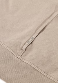 Beige Baumwoll-Sweatshirt mit feiner Textur, gerippten Bündchen und Saum, ausgestattet mit einem kleinen Stoffetikett an der Seite. Einfaches, minimalistisches Design.