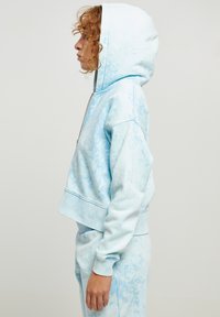 Lys blå croppet hettegenser med tie-dye mønster. Har justerbar hette og ribbestrikkede mansjetter. Laget av myk, teksturert stoff.