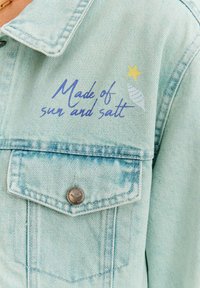 Jasnoniebieska kurtka dżinsowa z kieszenią na piersi, srebrnym guzikiem oraz nadrukowanym wzorem z tekstem "Zrobione z słońca i soli" i gwiazdką.