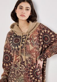 Sudadera con capucha de color beige que presenta intrincados patrones circulares y florales en burdeos, verde y crema. Textura suave, corte oversized.