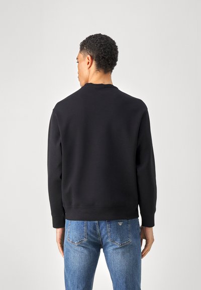 Felpa nera girocollo con maniche lunghe, caratterizzata da una texture liscia e un orlo a coste. Indossata con jeans in denim blu.