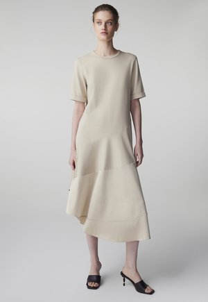 Jeune femme portant une robe midi asymétrique beige à manches courtes et des escarpins noirs à bout ouvert, debout devant un fond clair uni.