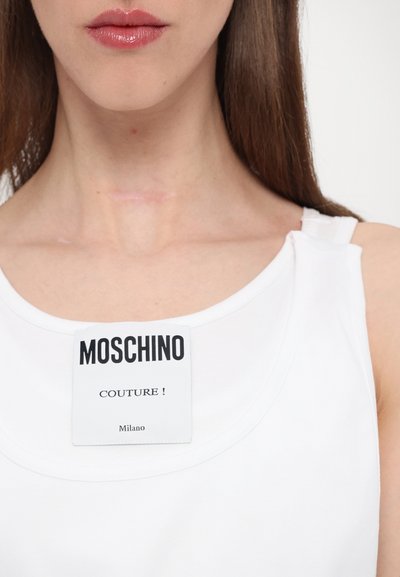Top alb fără mâneci, cu un decolteu rotund, având un eticheta proeminentă alb-negru care spune "MOSCHINO COUTURE! Milano."