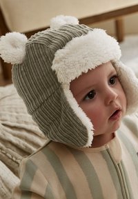 Zelená rebrovaná čiapka s bielym umelým kožúškom na okraji a medvedími uškami, ktorú nosí dieťa v pruhovanom béžovo-zelenom oblečení.