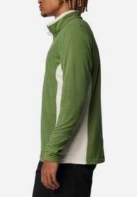 Uomo in giacca in pile verde e bianca con colletto alto, in piedi di lato, indossando pantaloni neri su sfondo bianco puro.