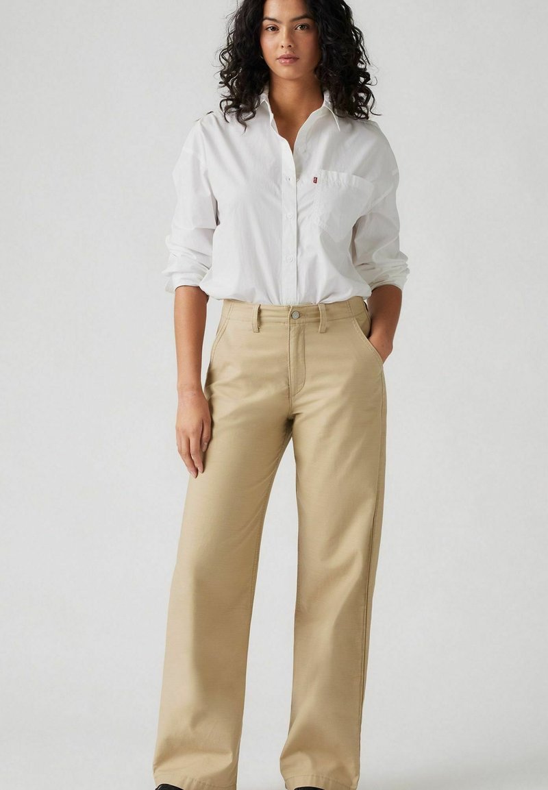 Pantalon large beige en tissu lisse, avec une fermeture à bouton sur le devant et des passants de ceinture, associé à une chemise blanche à col.