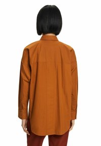 Camisa de manga larga, con botones, en tela de algodón de color óxido, con cuello puntiagudo y bajo curvado, y puños con botones visibles.