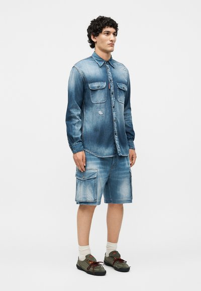 Άνδρας όρθιος, φοράει ξεθωριασμένο μπλε τζιν πουκάμισο και αντίστοιχα cargo σορτς, λευκές κάλτσες και ελιά πράσινα αθλητικά, σε απλό φόντο.