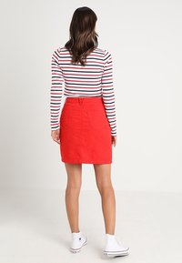 Jupe en denim rouge avec deux poches arrière, portée avec un haut à manches longues rayé. Des baskets blanches complètent la tenue.