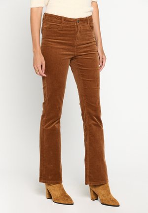 Bruine corduroy broek gedragen door een persoon met beige suède laarzen met hak en een wit textuur top, staand tegen een effen lichte achtergrond.