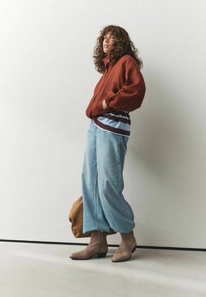 Femme aux cheveux bouclés portant une veste couleur rouille, une chemise rayée, un jean bleu large et des bottines beiges, debout contre un mur uni.