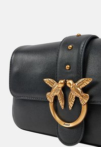 Sac en cuir noir avec un fermoir en forme d'oiseau doré, accent rond et clous décoratifs. Texture lisse et forme structurée.