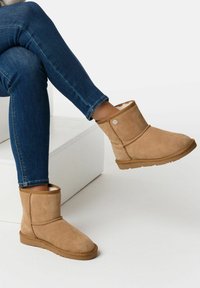 Tan-farbene Wildleder-Stiefeletten mit einem weichen Innenfutter, ausgestattet mit einem dekorativen Knopf an der Seite und flachen Gummisohlen. Tragen Sie sie mit blauen Jeans.