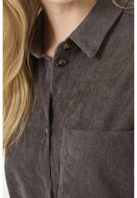 Fløjlsnederdel i mørkebrun med en krave med knapper, der har to brune knapper og en brystlomme. Tekstureret stof med lodrette striber.