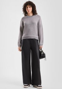 Grauer gestrickter Pullover mit geripptem Saum und weißem geometrischem Muster. Schwarze Hosen mit weitem Bein, glänzender Textur und Plateausneakern. Kleine Tasche.