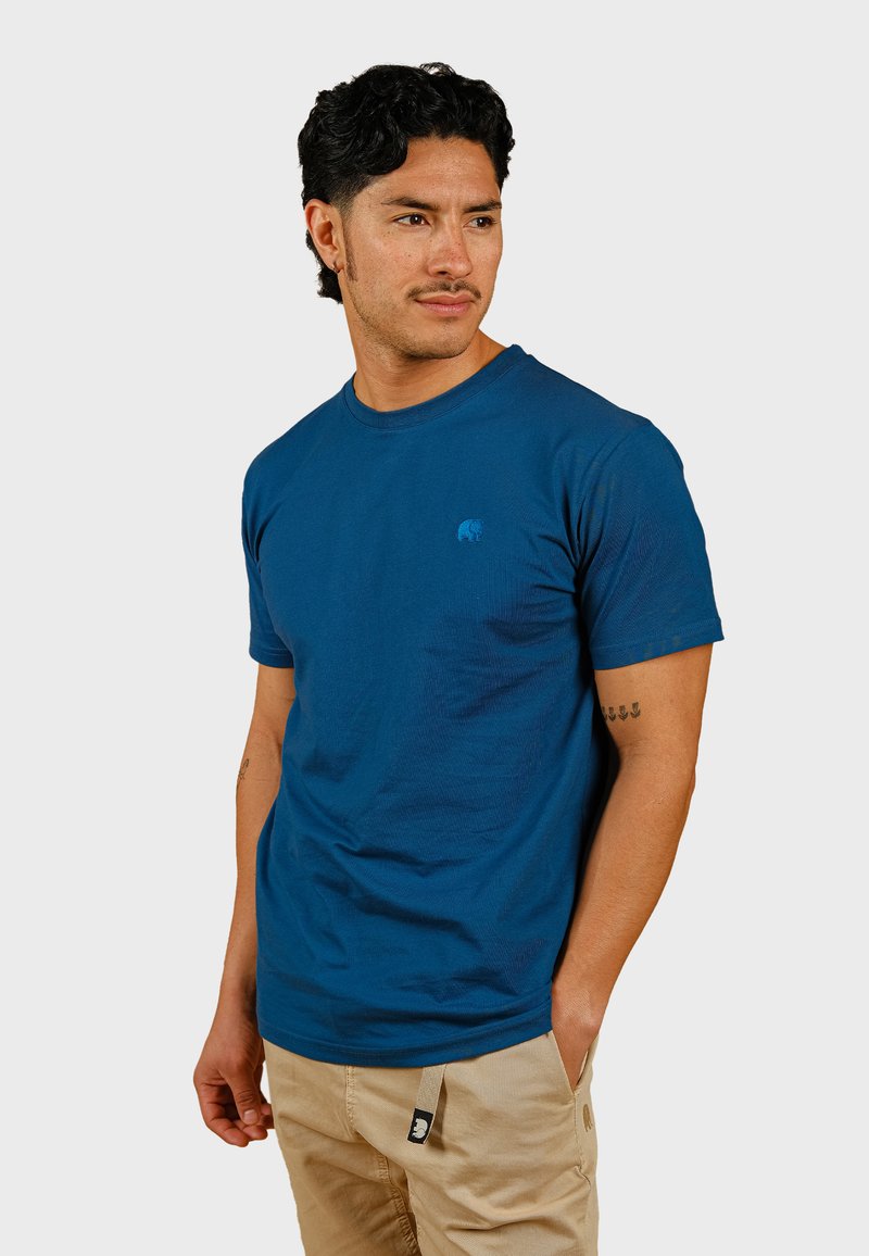 Camiseta de algodón azul con escote redondo y mangas cortas, con un pequeño y sutil logo en el pecho. Pantalones beige visibles.