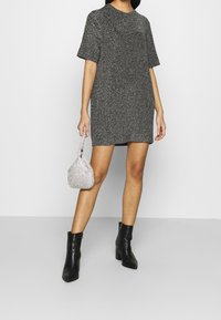 Vestido corto negro brillante de manga corta con un corte relajado, acompañado de un pequeño bolso estructurado plateado y botas negras hasta el tobillo.