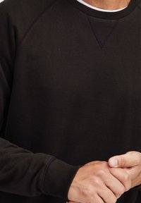 Sweat-shirt marron foncé avec un col et des poignets côtelés, une texture lisse et un design de manches raglan. Les mains sont jointes.